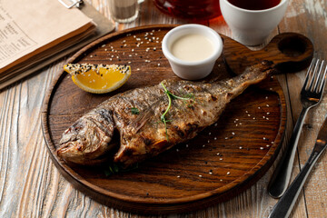 Whole grilled freshwater fish with head, garnished with sour cream and lemon, top view of rustic dinner, wooden background