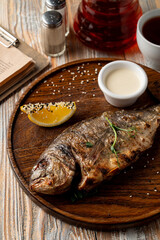 Whole grilled freshwater fish with head, garnished with sour cream and lemon, top view of rustic dinner, wooden background