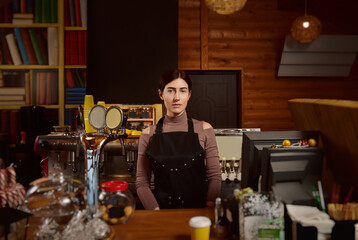 Portrait of a cute brunette woman barista in apron standing behind the bar
