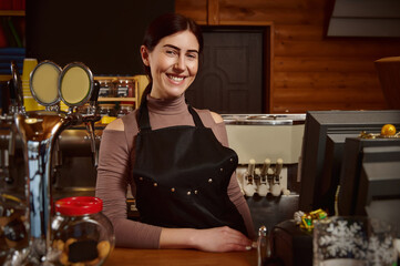 Young attractive smiling barista in apron standing behind the bar