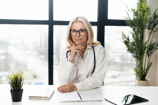 An Intelligent Mature Female Woman Doctor With Blonde Hair In A White Medical Robe, Glasses, And Stethoscope Sitting At The Desk With Papers, Documents, Notes, And Green Plants, Looking At The Camera