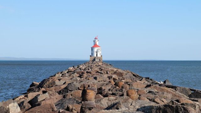 Lighthouse with red roof at the end of a pier on Lake Superior at Wisonsin Point. Large boulders surrounded by ripples of blue water and blue sky on a sunny day. Slow motion clip..