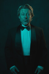 Fototapeta premium Blonde man wearing tuxedo and bow tie. Lit by blue and orange light against dark wall.