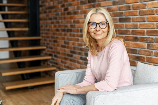 Portrait of a cheerful mature blonde woman sitting on the chair and smiling against a brick wall background. A successful middle-aged woman in glasses wearing casual clothing and looking at the camera