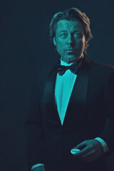 Man in tuxedo and bow tie holding casino dealer coin. Lit by blue and orange light in front of a dark wall.