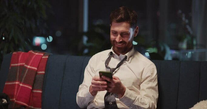 Attractive Millennial Businessman With Loosen Necktie Arriving Back Home Using Smartphone Social Media Rejoicing Leisure Relaxing Evening After Work.