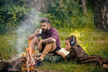 Couple relax on camp at bonfire in forest. Man and woman at campfire. Hipster drink and girl read book, listen to music. Summer vacation concept. Camping, hiking, lifestyle