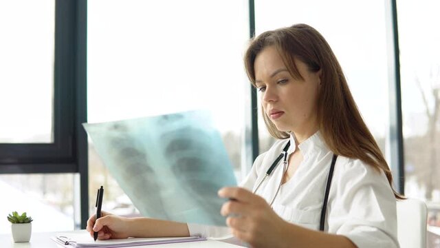 Young caucasian woman doctor wearing white medical coat and stethoscope writes a medical history in a notebook and holds the x-ray