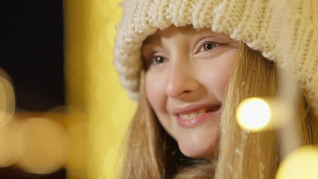 Close-up face of smiling cute Caucasian girl looking away with golden Christmas lights around. Portrait of happy child enjoying New Year night in city outdoors.