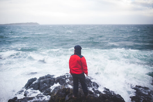 Man in red rain jacket standing on the ocean's shore getting hit by stormy waves