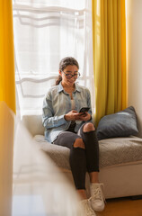Smiling brunette young woman sitting on couch and using smart phone in her room
