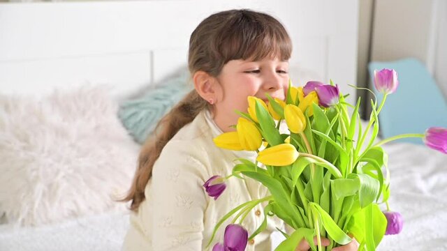 Laughing little girl with a bouquet of bright tulips in her hands, she sniffs them and smiles. A gift for mom or grandmother on women's day. 8 March Women's Day concept. High quality FullHD footage