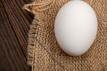 A chicken egg and a sewing needle on a homespun fabric with a rough texture.