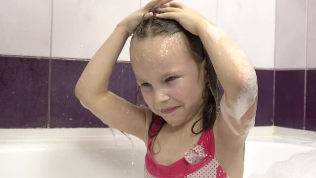 A little girl washes off the lathering shampoo with a shower.She sits in a bubble bath in a pink swimsuit.Squinting eyes from the pouring water.