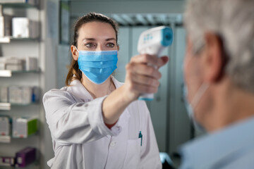 Fototapeta na wymiar Cinematic shot of female pharmacist with medical mask is measuring temperature with infrared thermometer gun to male patient in pharmacy. Concept of covid-19, protection, virus transmission, safety.