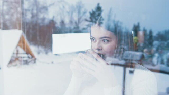 Young caucasian woman drinking hot beverage and looking through the window at snow covered houses. High quality 4k footage