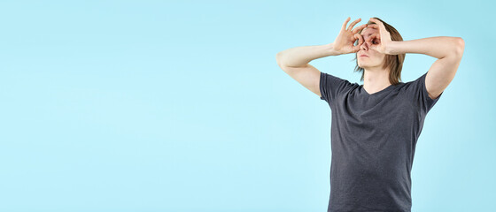 An emotional young man shows gestures. A portrait of a funny guy in a T-shirt isolated on a blue background makes an ok gesture similar to binoculars, eyes looking through fingers.