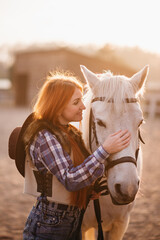Obraz premium A young cowgirl woman stroking a horse in the sunset at her ranch. Flare effect.