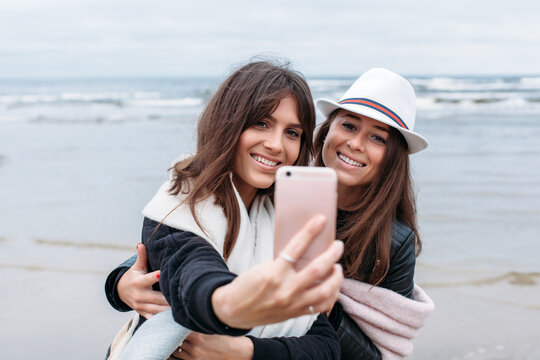 Two happy women fashion sensual style have fun together on the beach in cold autumn wether, warm dressed posing while doing selfie photos on smartphone camera