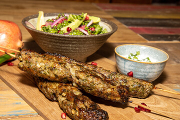 A delicious pile of three Spiced Lamb Kaftas kababs with a bowl of salad and pomegranate seeds on a wooden kitchen work top