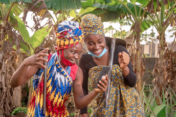 Two beautiful African female farmers with nose mask, looking happily into a smartphone and holding hoe and cutlass in a banana plantation  