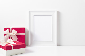 White blank photo frame, two pink gifts with white satin bow on white table. Front view. Place for text, copy space, mockup