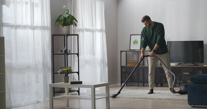 adult handsome man is hoovering carpet in apartment, using vacuum cleaner for cleaning room at weekend