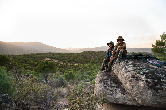 Two boys sitting on a stone covered with a blanket watching the sunset
