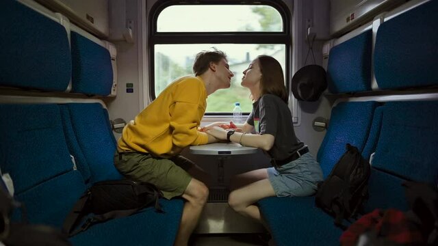 Positive young couple sitting at a table in a train holding hands, talking with smiles on their faces and kissing. Couple travels by train.