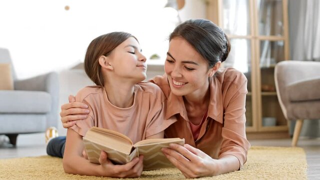 Lockdown of Caucasian teenage girl and young mixed-race woman lying in embrace on carpet in living room, reading book and then looking at camera