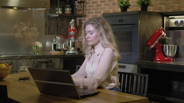 Beautiful girl is sitting in kitchen evening drinking coffee and looking at  laptop screen. woman is engaged in successful business development remotely. Freelancer works online computer in evening