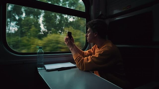 Young man hiking in orange sweater walks in train and makes landscape photo from window on smartphone camera. Railway travel concept