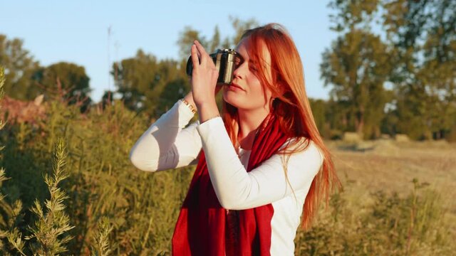[4k] slow motion portrait of beautiful ginger woman taking a picture with vintage slr outdoors in nature at war sunset