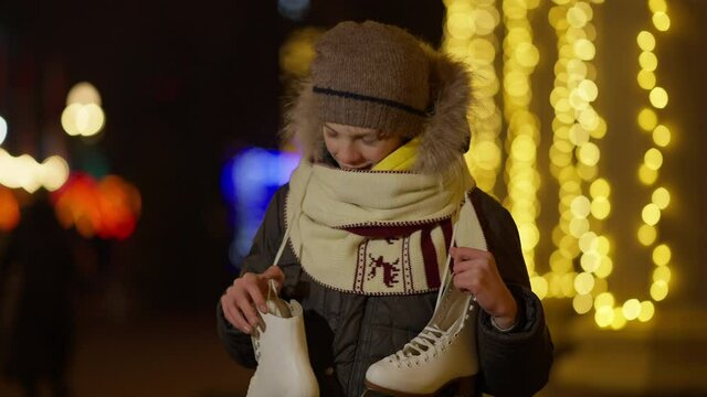 Happy Caucasian boy boasting new ice skates standing on New Year in night city with golden Christmas lights at background. portrait of excited kid bragging holiday gift.