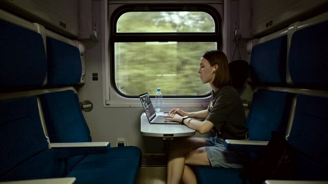 Beautiful young woman in casual clothes working on a laptop at a table in a train and communicates on wireless headphones. Female freelancer working on laptop while traveling by train.