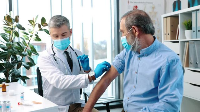 Close up of middle aged handsome man physician doctor in medical mask making vaccine injection to Caucasian senior patient sitting in hospital cabinet lab, coronavirus vaccination concept