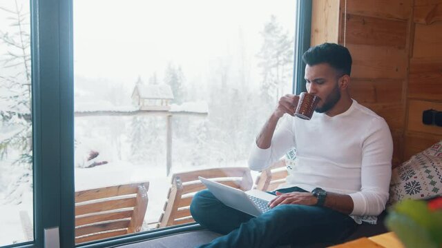 Young man browsing social media and drinking tea near the window on winter vacation. High quality 4k footage