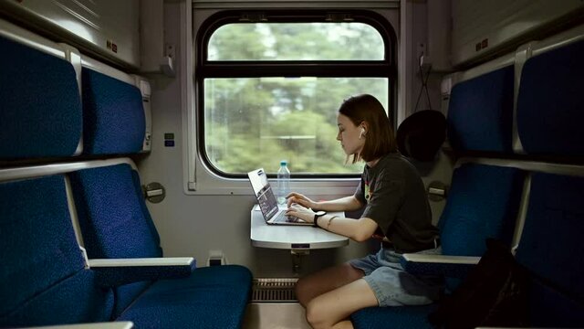 Attractive woman in wireless headphones rides a train and works on a laptop in a compartment at a table while moving. Work on the Internet while traveling.