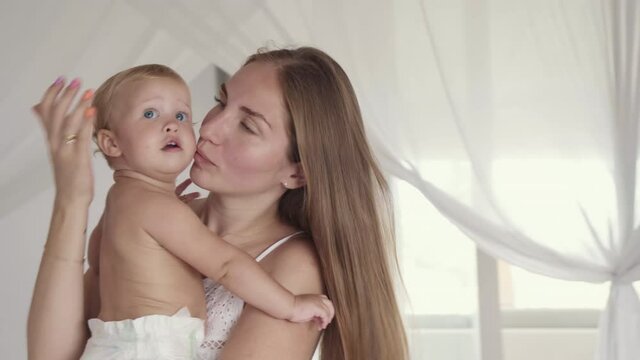 Medium close-up of young long-haired Caucasian mother holding her blue-eyed one-year-old baby wearing diaper. Woman talking to, hugging, kissing toddler