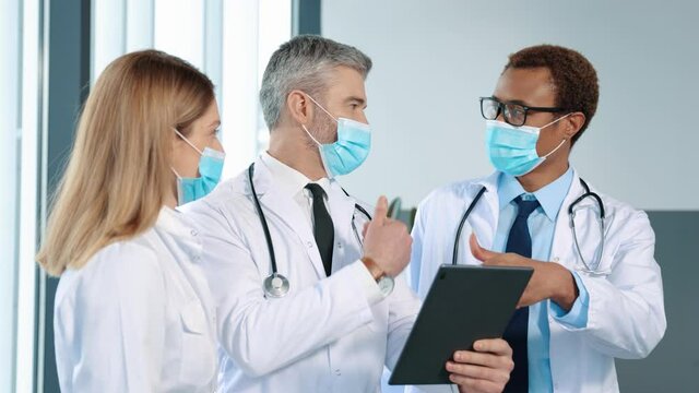 Male head of clinic standing with team of mixed-races doctors colleagues and using tablet device while discussing something. Group of multi ethnic medics, men and women talking and having discussion