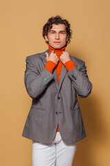 Tall handsome man dressed in orange turtleneck, white jeans and grey jacket posing on the beige background