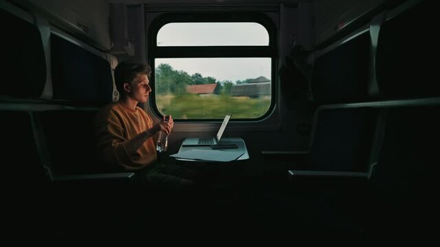 Handsome young man working on laptop in train at table in dark compartment and drinking water from bottle. Freelancer working on laptop and internet on the go while the train is moving