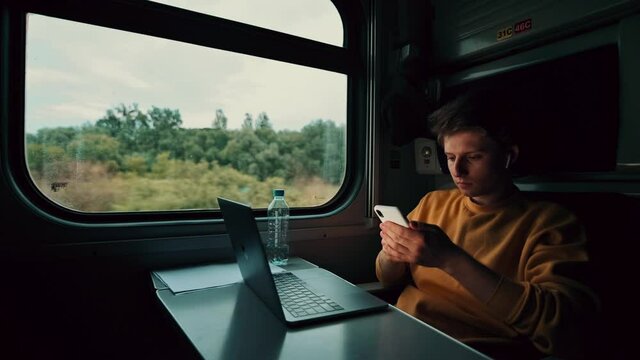 Young man in wireless headphones sits while traveling by train at a table with a laptop and uses a smartphone with a serious face.