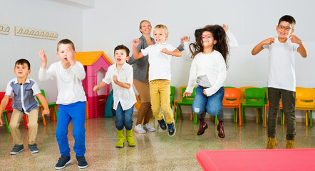 Positive happy cheerful smiling female teacher and group of schoolkids jumping in classroom