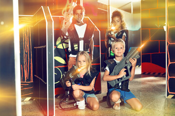Cheerful tween girls and boys of different nationalities with laser pistols posing together at a dark laser tag labyrinth