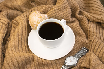 A white cup of coffee with a gingerbread on a saucer sits on a warm sweater next to a wristwatch.