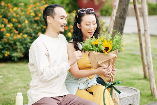 Happy excited young Chinese woman looking at beautiful bouquet from boyfriend when they are sitting on bench in park