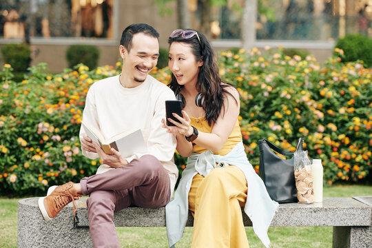 Young Chinese man laughing at post or photo on social media that his girlfriend is showing when they are sitting on bench outdoors