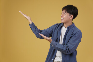 portrait of happy handsome young asian man dressed casually presenting with open palms gesture at copyspace. yellow background
