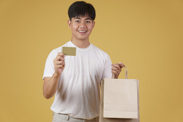 portrait of happy young asian man dressed casually holding shopping bags and credit card isolated on yellow background